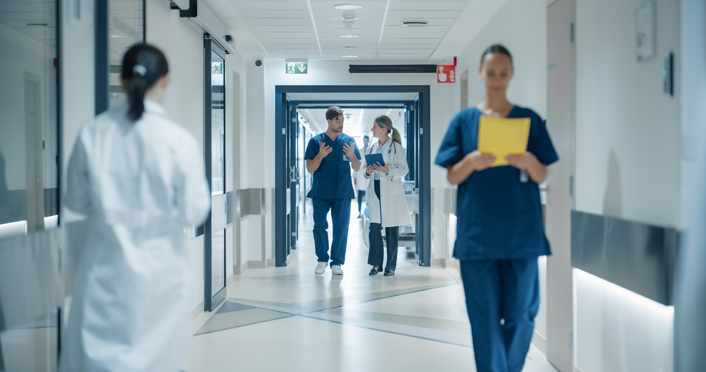 Healthcare Professionals Walking to Their Examination Rooms, Reception Desk, ICU Units in a Modern Bright Hospital Corridor. Female Doctor Showing a Young Male Surgeon Information on a Tablet